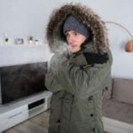 Person wearing a warm parka with a fur-lined hood, looking cold indoors, emphasizing the need for effective heating and plumbing services.