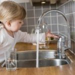 Child reaching for a kitchen faucet in a modern setting with gray tiles and wooden countertops.