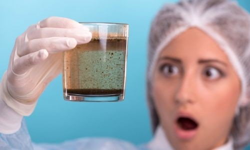 Woman in protective gear holding a glass of yellowing water, expressing shock at its appearance, highlighting plumbing issues related to water quality.