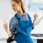 Woman in blue overalls holding a plunger and plumbing pipe, representing female participation in the plumbing industry and the evolving image of plumbers.