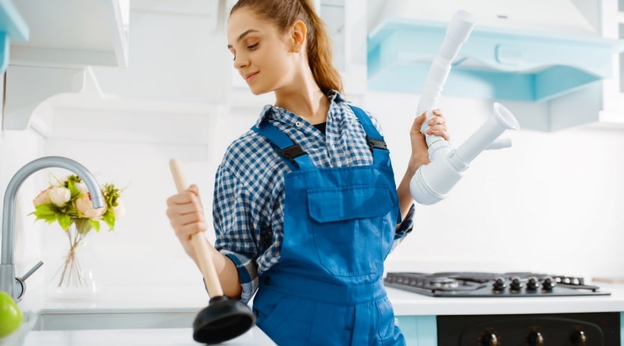 Woman in blue overalls holding plumbing tools and a plunger in a modern kitchen, representing Marc Francis Plumbing services.