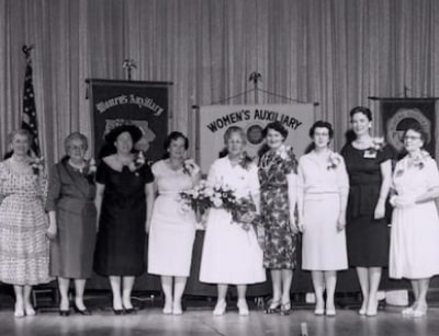 Women of the Ladies Auxiliary Committee for the National Association of Plumbers standing together at an event, showcasing their activism and contributions to plumbing history, with banners in the background.