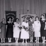 Women in formal attire standing on stage with floral arrangements, celebrating contributions to the plumbing industry, alongside banners for the Women's Auxiliary Committee.