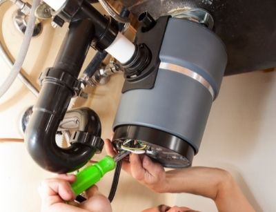 Person repairing a garbage disposal under a kitchen sink, emphasizing plumbing maintenance for holiday meal preparation.