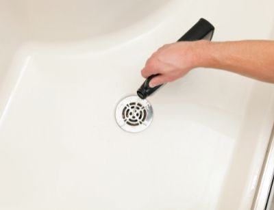 Hand holding a drain cleaning tool above a shower drain, illustrating the dangers of using liquid drain cleaners for plumbing maintenance.