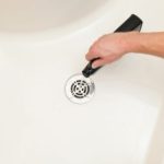 Hand using a drain cleaning tool above a shiny drain in a clean bathtub, highlighting plumbing maintenance for homeowners.