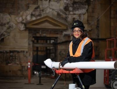 Adrienne Bennett, wearing a construction helmet and safety vest, smiling while holding a rolled-up blueprint in a plumbing worksite, symbolizing her achievements as the first African-American female master plumber in the U.S.