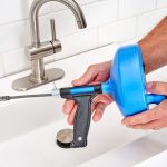 Person using a blue drain auger to unclog a white sink with a stainless steel faucet in a clean workspace.