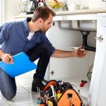 Man kneeling at a kitchen sink for plumbing repairs, toolbox nearby, illustrating services by Marc Francis Plumbing.