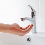 Hand reaching for a modern chrome faucet with dripping water, representing cleanliness for homeowners wary of frozen pipes.