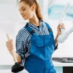 A young woman in blue overalls holds a plunger and plumbing pipe, showcasing women's roles in plumbing services.