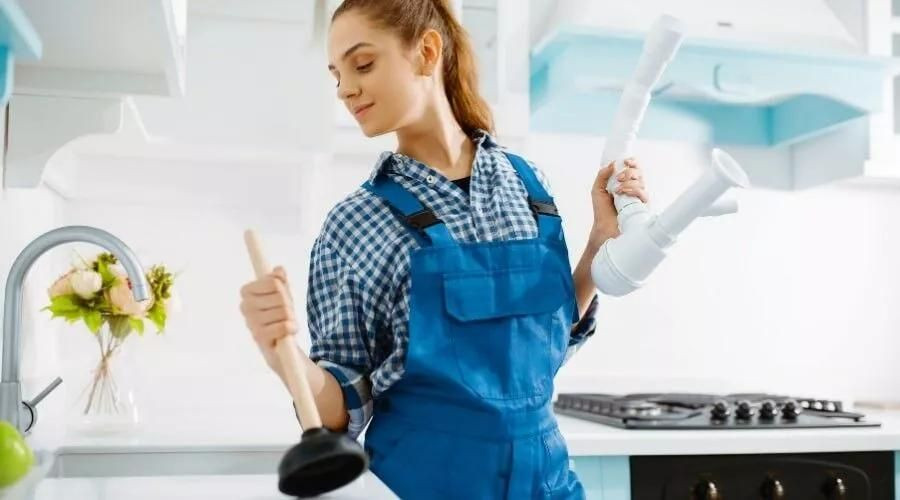 Woman in blue overalls holding a plunger and plumbing pipe in a bright kitchen, representing plumbing solutions and maintenance.