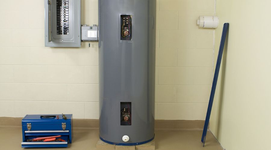 Gray water heater cylinder with gauges and valves, toolbox, and electrical panel in organized plumbing maintenance space.