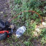 Gardening tools on soil and grass, including a hose and bag, for maintenance in outdoor plumbing areas.