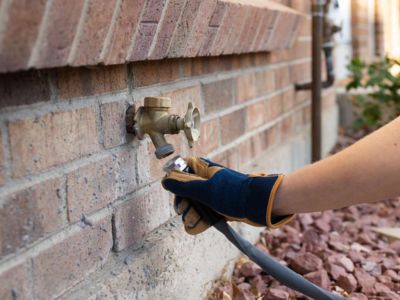 Person wearing gloves disconnecting a garden hose from an outdoor faucet on a brick wall, illustrating winterization of outdoor plumbing to prevent freezing pipes.