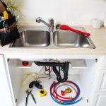 Kitchen sink with plumbing tools, including a red plumber's wrench, under-sink pipes, and various plumbing supplies, emphasizing plumbing maintenance and repair.